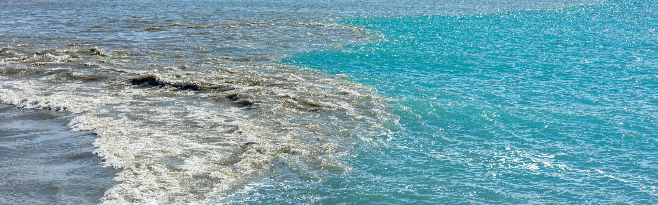 Clean, turquoise sea water near a beach. From the left dark, murky sewage or dirty runoff water is merging creating a dirty mixture between the contrasting blue ocean