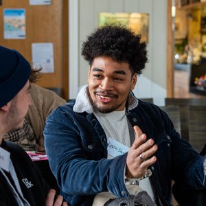 A young man with dark curly hair is wearing a blue corduroy jacket with a white fleece collar, he is smiling and gesturing with his hands while talking to another person. The person he's speaking to is wearing a blue beanie and a black hoodie.