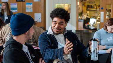 A young man with dark curly hair is wearing a blue corduroy jacket with a white fleece collar, he is smiling and gesturing with his hands while talking to another person. The person he's speaking to is wearing a blue beanie and a black hoodie.