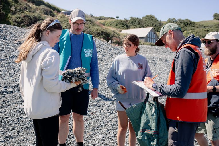 Marine Conservation Society staff working with young people on a beach. One girl is holding a large litter item, while a staff member fills out the litter survey paper form.