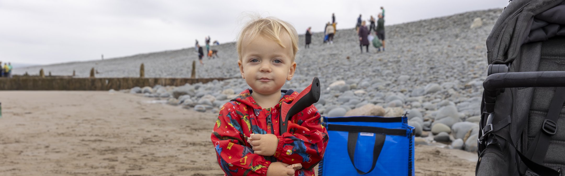 A child wearing a red dinosaur patterned outfit and blue wellies is holding a litter picker and posing for the camera