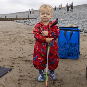 A child wearing a red dinosaur patterned outfit and blue wellies is holding a litter picker and posing for the camera