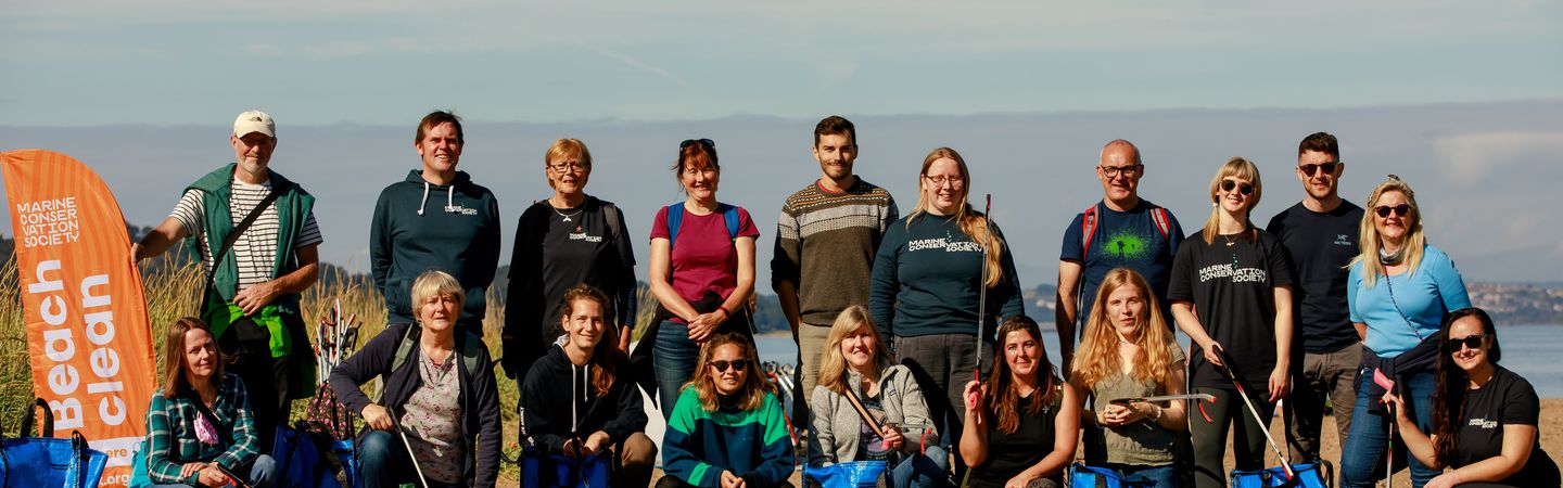 A group of 18 volunteers, some standing and some crouching, pose on a sandy beach with blue collection bags and litter pickers after a beach clean event as part of the Marine Conservation Society's Beachwatch program