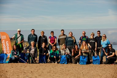 A group of 18 volunteers, some standing and some crouching, pose on a sandy beach with blue collection bags and litter pickers after a beach clean event as part of the Marine Conservation Society's Beachwatch program