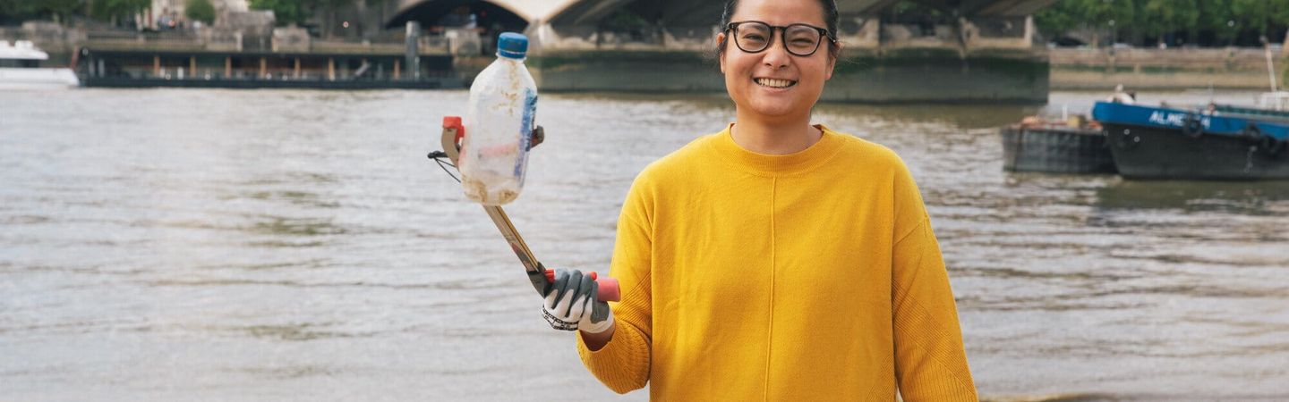 A woman in a yellow jumper holding up a plastic bottle to the camera using a litter picker. She is in front of the river at Festival Pier, London