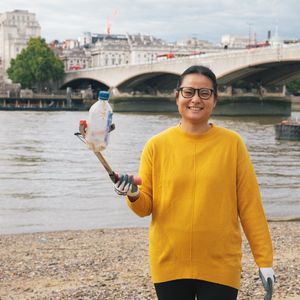 A woman in a yellow jumper holding up a plastic bottle to the camera using a litter picker. She is in front of the river at Festival Pier, London