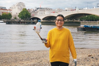 A woman in a yellow jumper holding up a plastic bottle to the camera using a litter picker. She is in front of the river at Festival Pier, London
