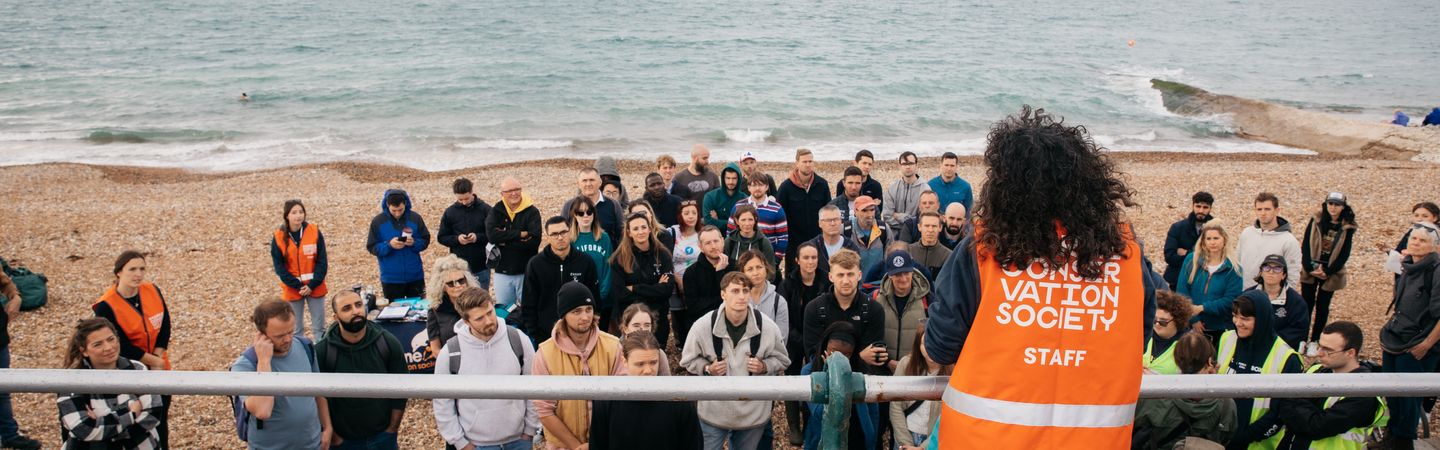 A woman with her back to the camera speaking to a group of beach cleaners all huddled in front of her on the beach. The woman is wearing a Marine Conservation Society vest.