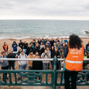 A woman with her back to the camera speaking to a group of beach cleaners all huddled in front of her on the beach. The woman is wearing a Marine Conservation Society vest.