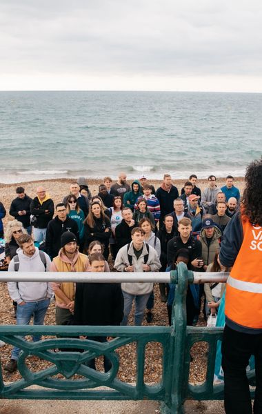 A woman with her back to the camera speaking to a group of beach cleaners all huddled in front of her on the beach. The woman is wearing a Marine Conservation Society vest.