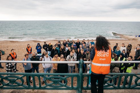 A woman with her back to the camera speaking to a group of beach cleaners all huddled in front of her on the beach. The woman is wearing a Marine Conservation Society vest.