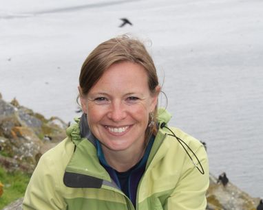 Miranda Krestovnikoff smiles at the camera from a rocky cliff face with the sea in the background. It appears there are puffins flying in the background, although their silhouettes are small.