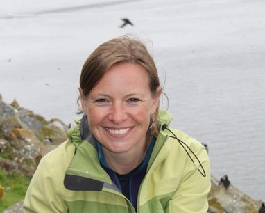 Miranda Krestovnikoff smiles at the camera from a rocky cliff face with the sea in the background. It appears there are puffins flying in the background, although their silhouettes are small.