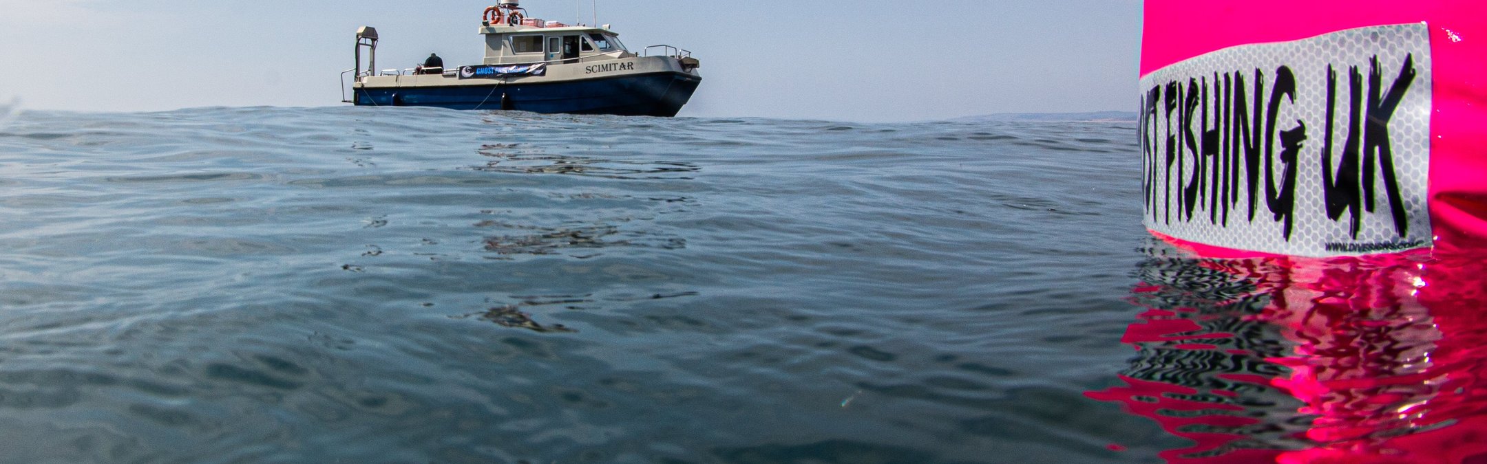 A fishing boat in shown in the distance with a bright pink buoy-type device reading 'ghost fishing UK' in the foreground.