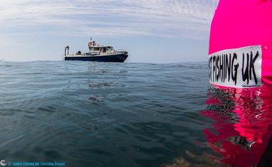 A fishing boat in shown in the distance with a bright pink buoy-type device reading 'ghost fishing UK' in the foreground.
