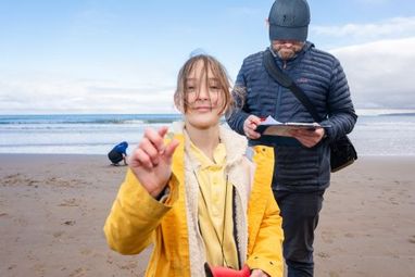 A young girl in a yellow coat holding up a stone. A teacher is standing behind her looking at a form. They are situated on a beach.